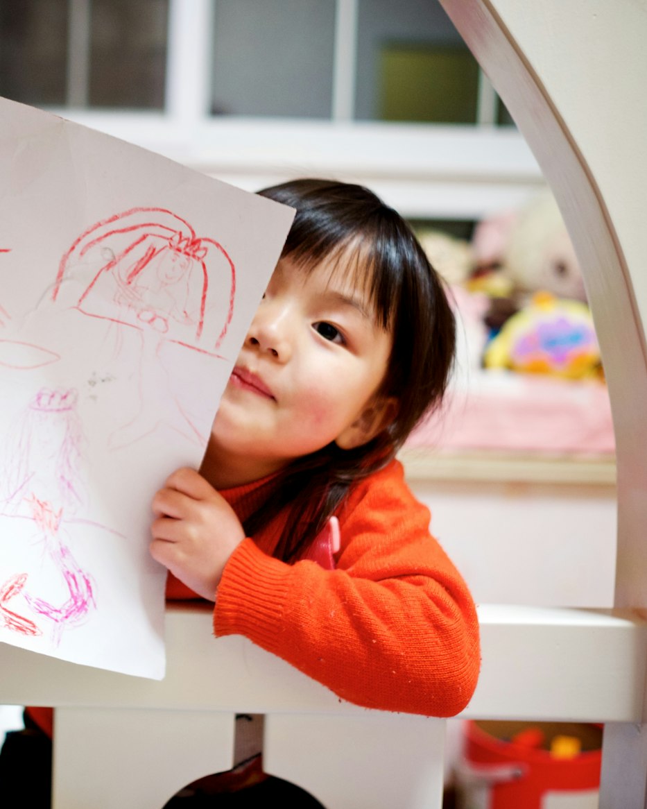 Une petite fille souriante montre son dessin au feutre rouge, dans une chambre claire