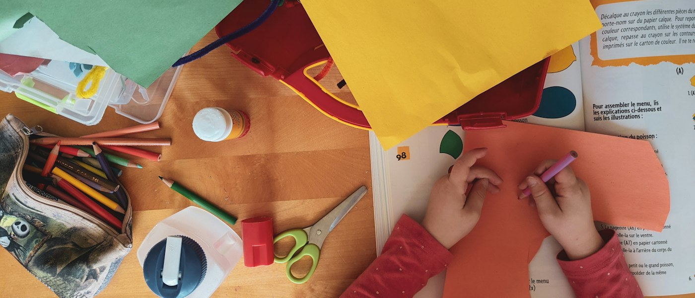 Vue de dessus : des mains d’enfant découpent du papier de couleur, entourées de feutres, de ciseaux et de colle