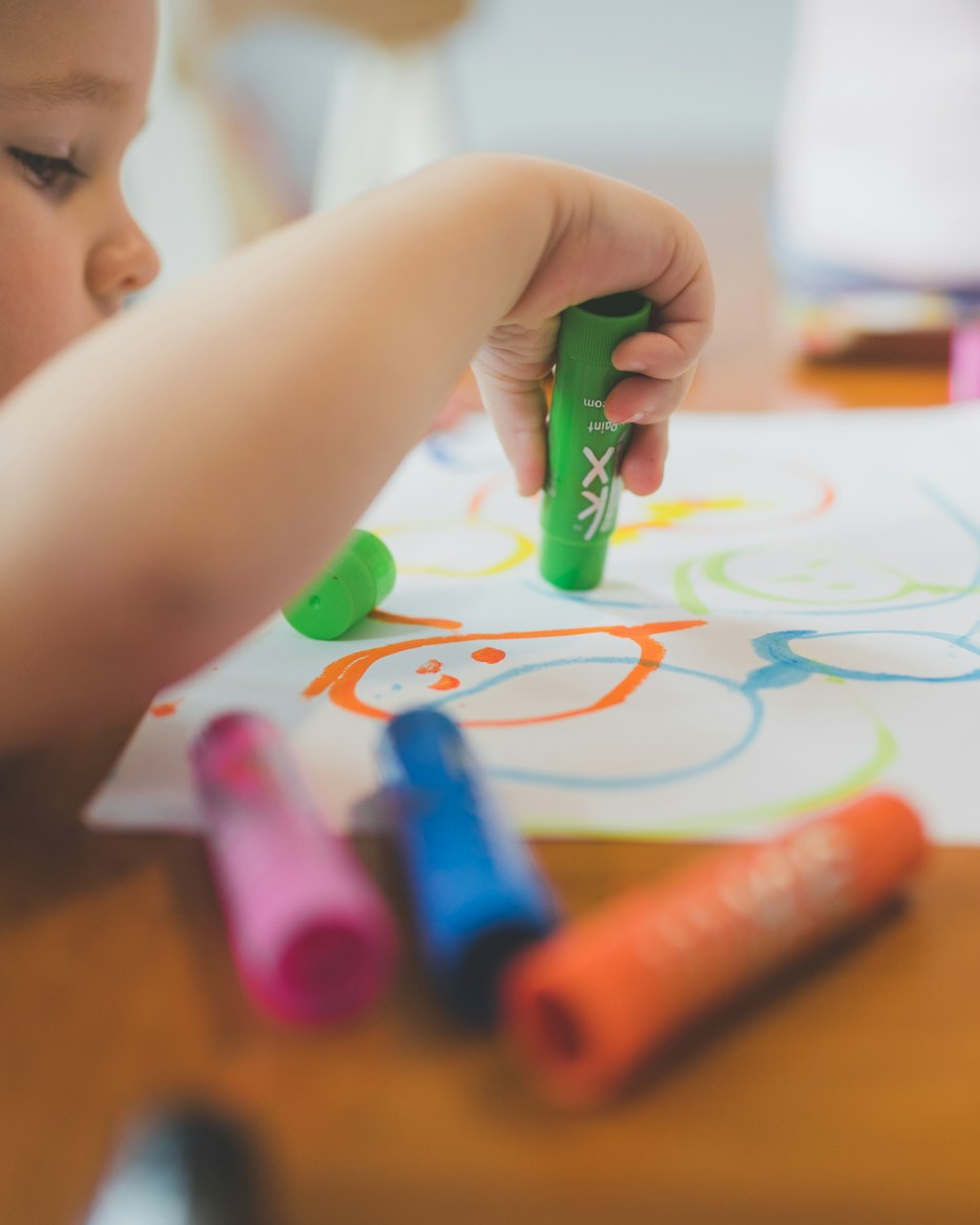 Un enfant dessine au feutre et à la craie grasse sur une feuille, sur une table en bois