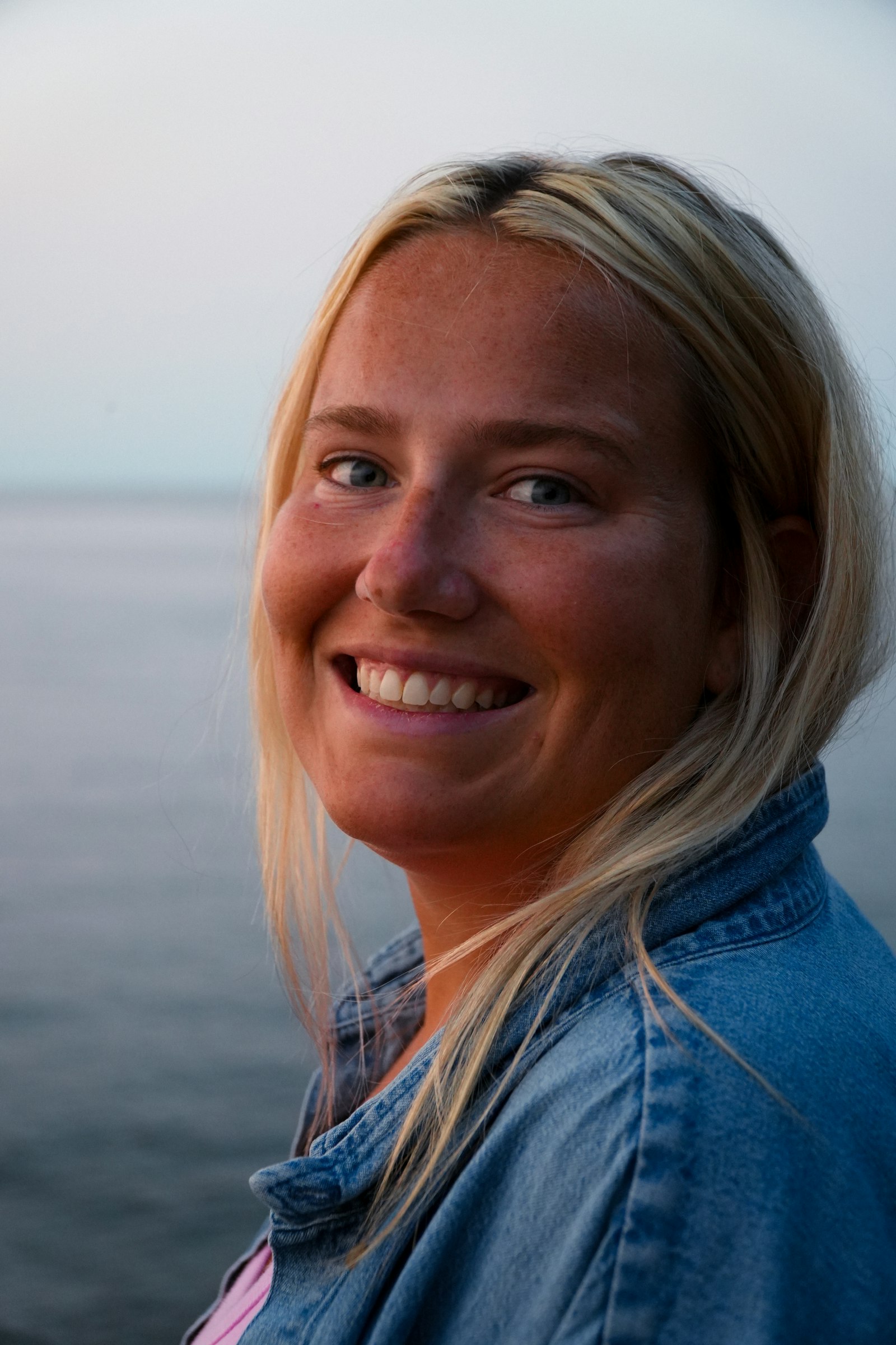 Une femme blonde souriante en veste en jean, tournée vers l’objectif, au bord de l’eau